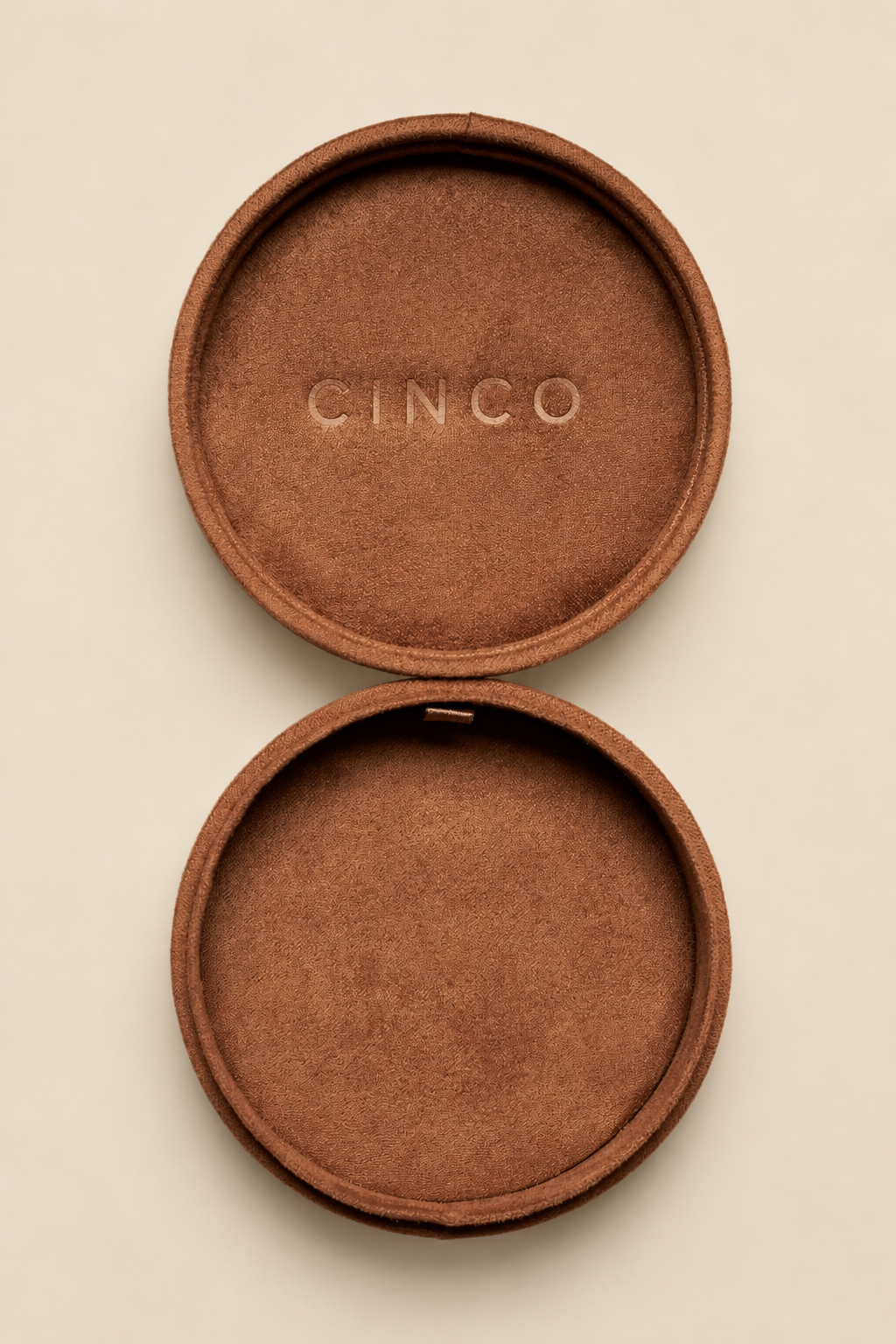 An open round makeup compact with a textured brown suede-like lid and base, featuring the brand name CINCO embossed on the top lid, set against a solid light beige background.