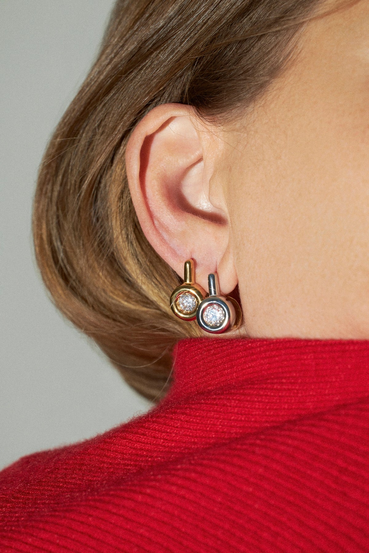 A close-up of a person's ear wearing two small hoop-style earrings with gold-toned posts and circular clear stone settings, paired with a bright red ribbed sweater against a light gray background.