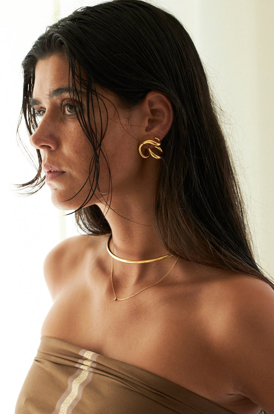 A woman in a taupe strapless top wearing gold jewelry, including a gold C-shaped earring and a delicate gold necklace, shown in profile against a bright, softly lit background.