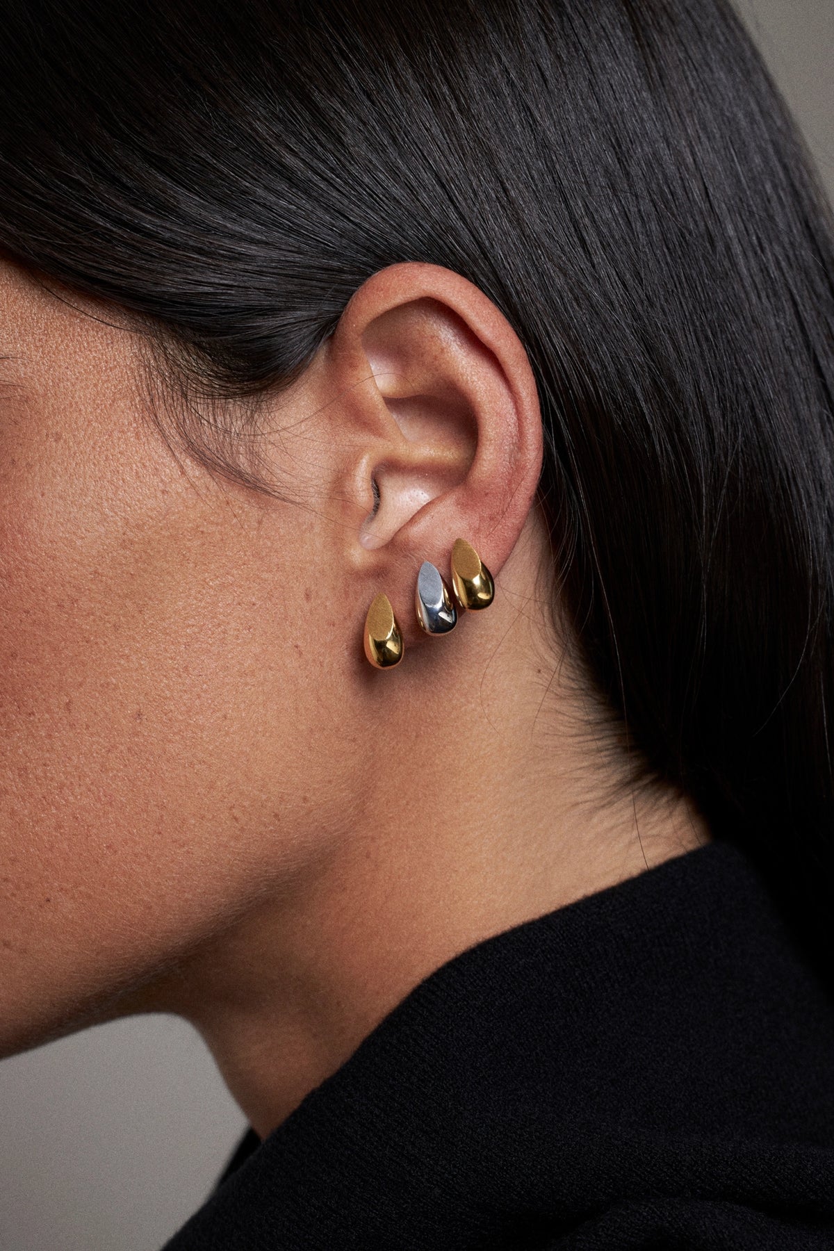 Close-up of a person's ear wearing three small teardrop-shaped earrings in gold, silver, and white, with dark hair and a black top visible.
