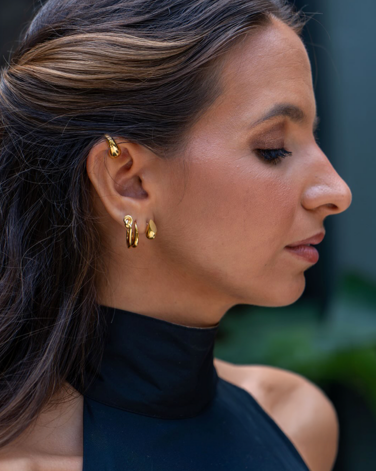 A close-up side profile of a woman wearing small gold hoop earrings and a gold ear cuff, with her dark hair and black high-neck top visible against a softly blurred dark background.