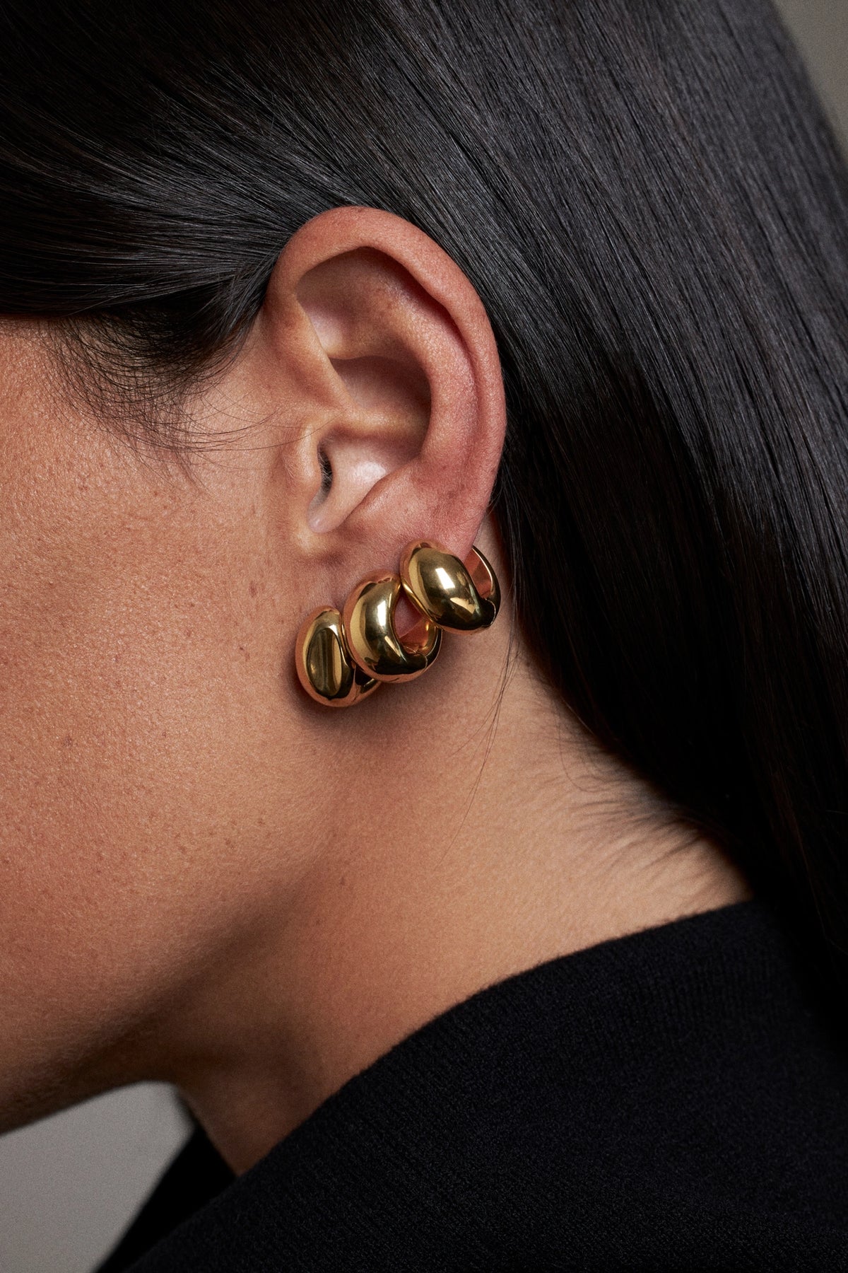 A close-up side profile of a person wearing a pair of gold hoop earrings with a textured, organic wavy design.