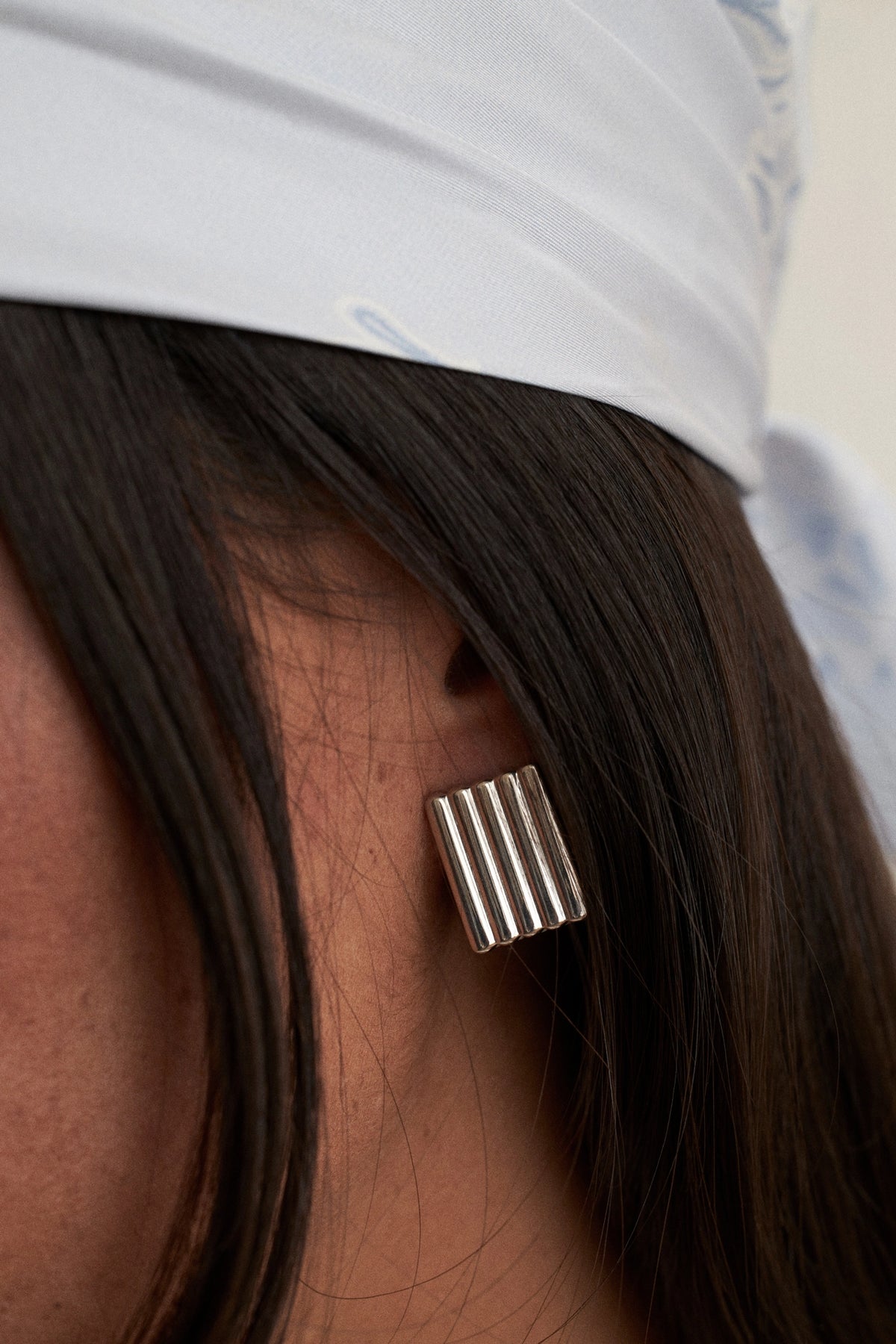Cannelé Rectangular Ribbed Earrings. A close-up of a person's ear wearing a silver rectangular stud earring with vertical grooved lines, next to dark hair and a white head covering. where can I find cool earrings online?