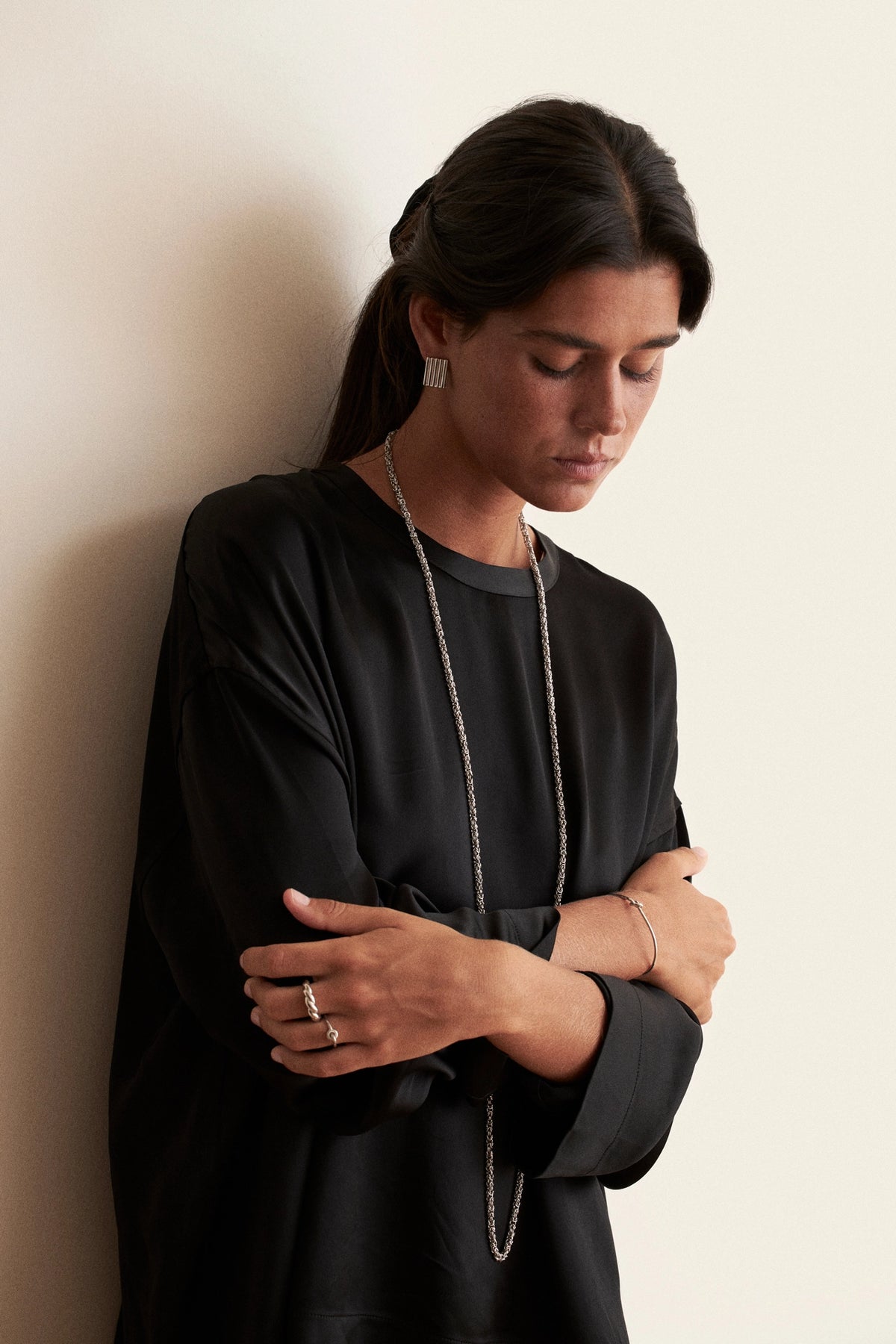 A woman with dark hair tied back wears a black long-sleeve top and a long silver chain necklace, standing against a plain off-white wall with her arms crossed.