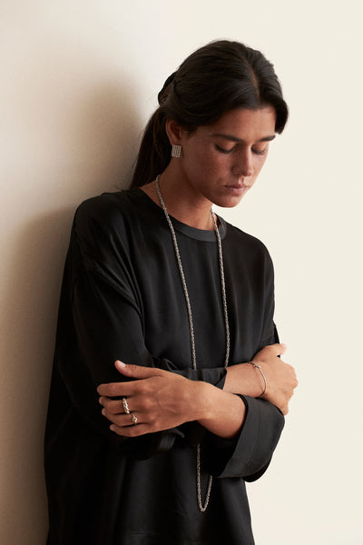 A woman with dark hair tied back wears a black long-sleeve top and a long silver chain necklace, standing against a plain off-white wall with her arms crossed.