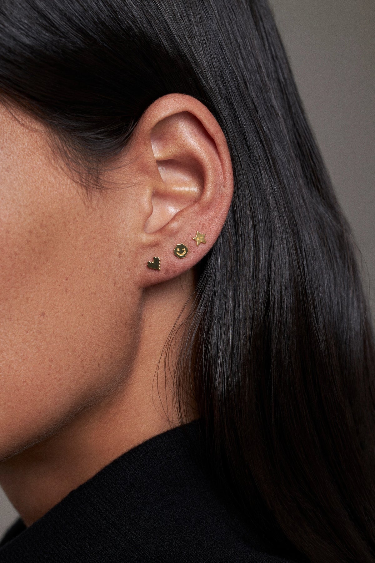 Close-up of a person's ear wearing three small gold and green stud earrings, with long dark hair and a black top against a neutral gray background.