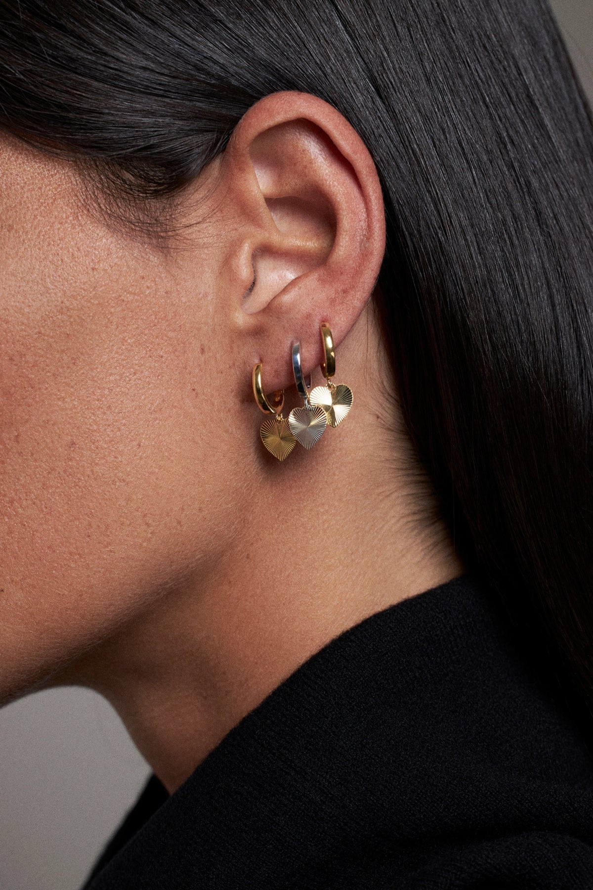 A close-up side profile of a person wearing a small gold hoop earring with dangling heart charms, one gold heart and one silver heart, against dark hair and a black top.